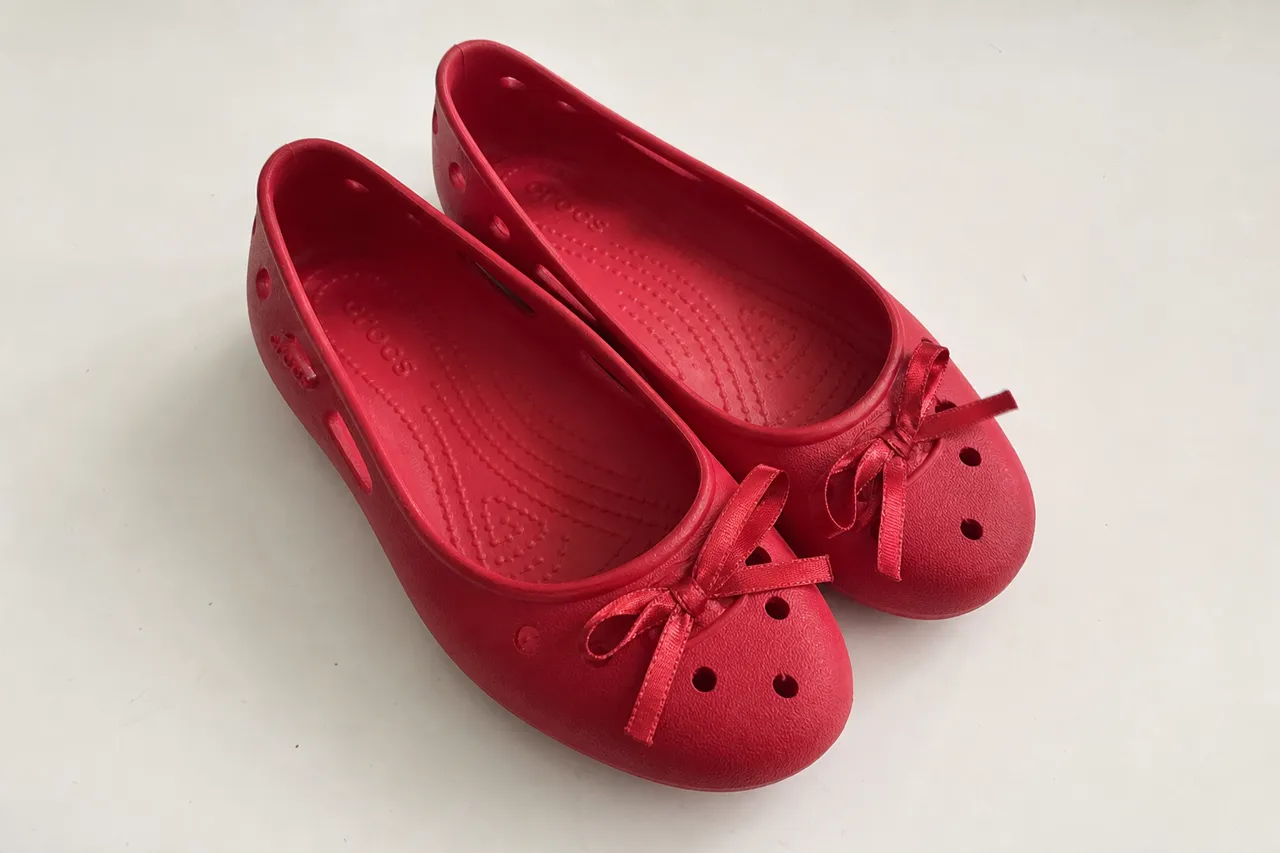 A close-up photograph of a pair of red Crocs Classic Ballet shoes with ribbon details, soft natural lighting on a minimalist white background, high-end fashion magazine style
