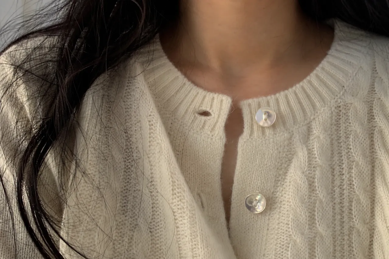 A close-up photograph of high-quality mother-of-pearl buttons on a cream-colored knit cardigan, focus on the shimmering texture of the buttons and delicate stitching, soft natural light
