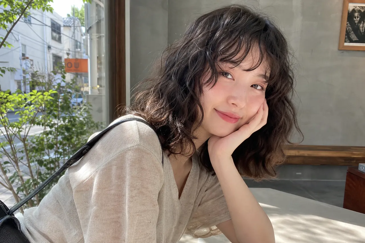 A stylish Korean woman with voluminous medium-length hippie perm and puppy bangs, looking chic in a summer cafe setting, natural sunlight, high-end photography style