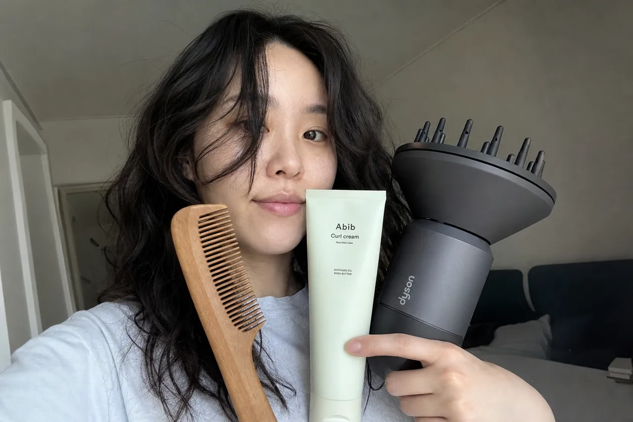 A collection of hair care products including a curl cream tube, a wooden comb, and a hair dryer with a diffuser nozzle, minimalist aesthetic, natural daylight
