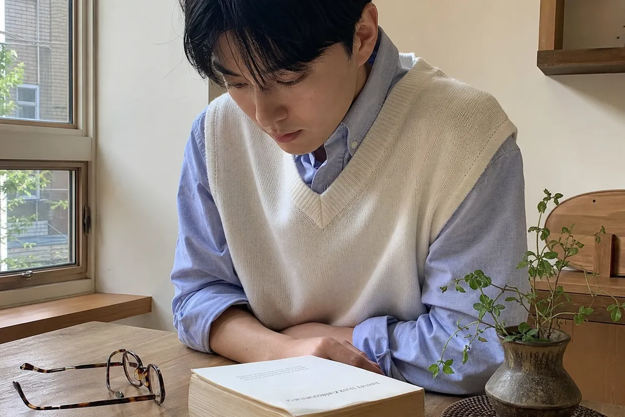 A warm-toned aesthetic photograph of a minimalist white vest and a light blue shirt layered on a wooden desk, with vintage glasses and a thick novel, soft morning sunlight, intellectual mood