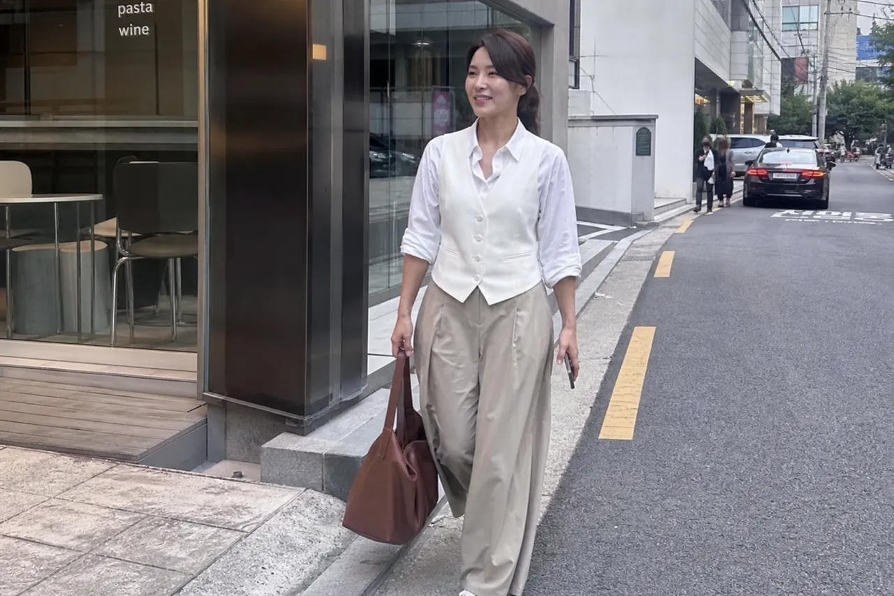 A full-body photograph of a stylish Korean woman in her 30s wearing a white vest layered over a shirt, wide-leg trousers, and carrying a large leather tote bag, walking on a modern city street, bright daylight