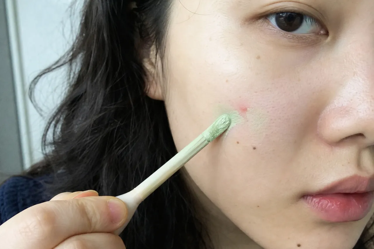 A close-up of a hand applying green color-correcting concealer on a red spot on skin, soft focus, high-quality beauty photography