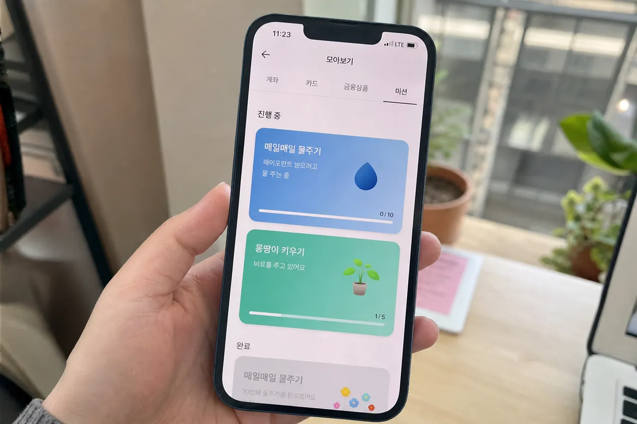 A close-up photograph of a smartphone screen displaying a banking app with colorful mission buttons for watering and fertilizer, soft natural indoor lighting