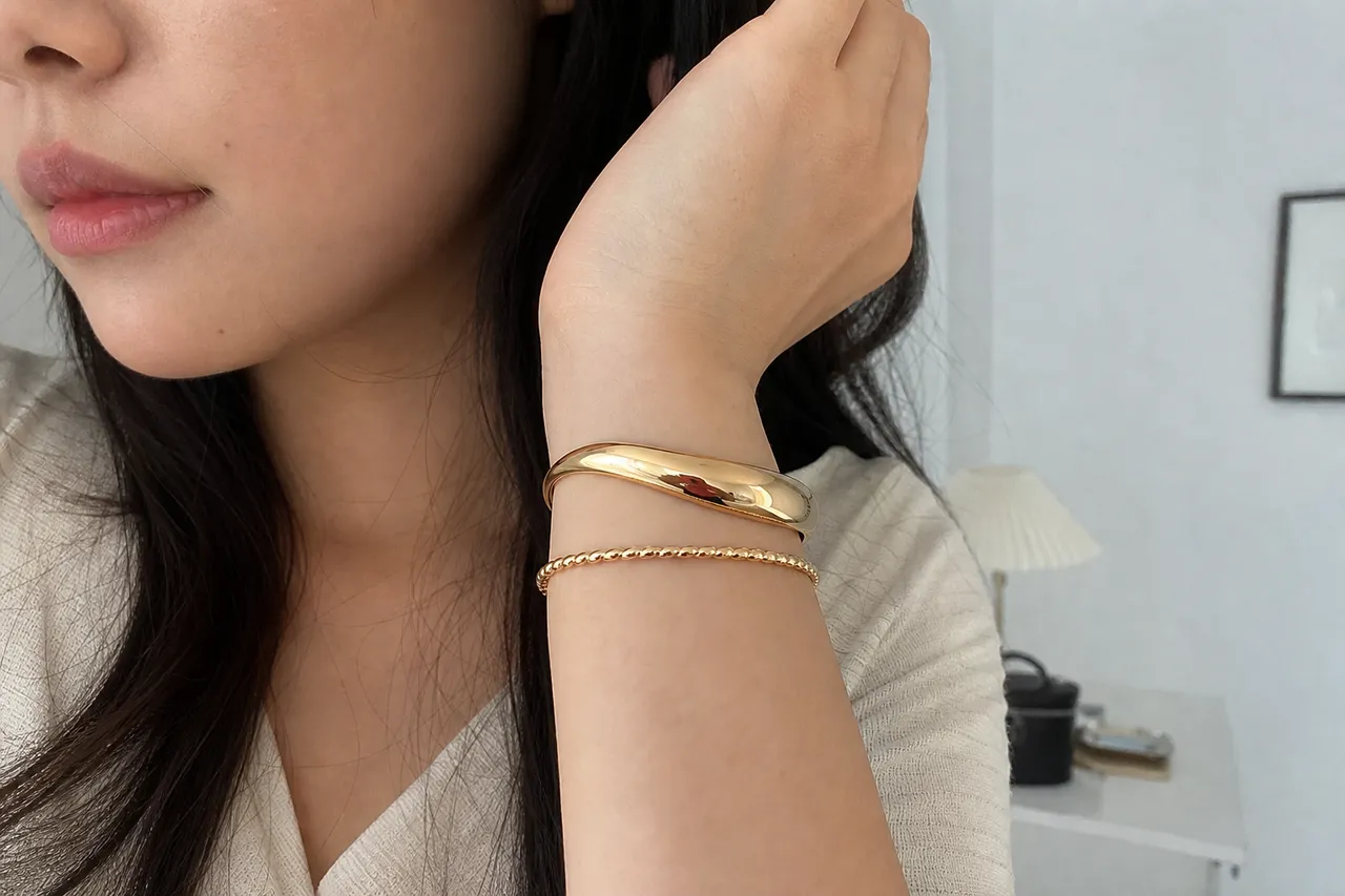 A close-up shot of two different gold bangles on a wrist, one slim and one bold, illustrating the layering technique, clean minimalist background, professional jewelry photography