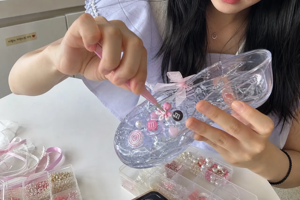 Close-up of a hand decorating a clear jelly shoe with cute charms and ribbons, DIY 'Shin-gu' trend, soft aesthetic lighting