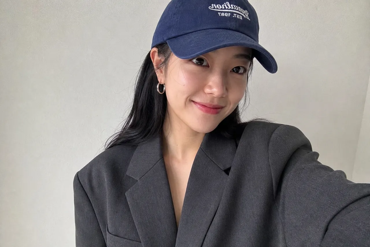 A close-up photograph of a classic charcoal grey oversized blazer paired with a casual navy blue baseball cap and silver hoop earrings, clean white background, studio lighting, minimal fashion aesthetic
