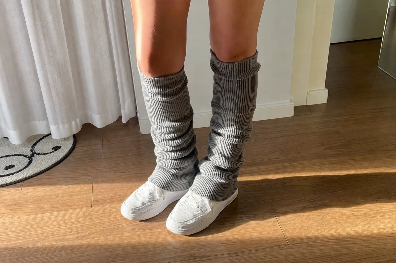 A close-up shot of a person's legs wearing grey ribbed leg warmers over white sneakers, standing on a wooden floor, soft morning sunlight, wellness and lifestyle vibe