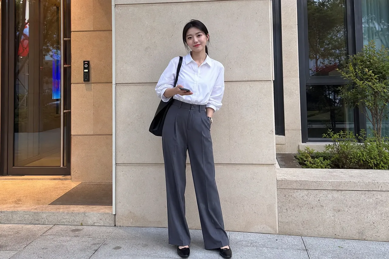 A full-body shot of a Korean woman wearing high-waisted wide-leg trousers and a tucked-in white shirt, standing against a clean architectural background, sharp fashion photography